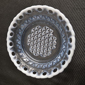 May include: A clear glass dish with a white, scalloped edge featuring evenly spaced holes. The center of the dish has a geometric pattern of triangles. The dish is round and sits on a dark surface.