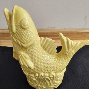 Puede incluir: Una escultura de pescado de cerámica amarilla con escamas y aletas detalladas. El pez está en posición vertical con la boca abierta y la cola curvada hacia un lado. La base de la escultura está decorada con plantas estilizadas.