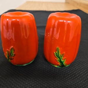May include: A pair of ceramic salt and pepper shakers. The shakers are a vibrant red color, shaped like a tomato, and feature a green leaf design. The shakers are on a black surface.