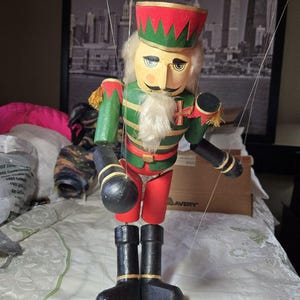 May include: A wooden nutcracker puppet with a red and green hat, green jacket, red pants, and black boots. The puppet has a white beard and mustache, and is suspended by strings. The background includes a cityscape.