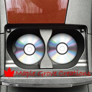 Puede incluir: Dos portavasos negros con ribete plateado, cada uno con un CD plateado y arcoíris. Los portavasos están en la consola central de un coche. El texto "Maple Grove Creations" está escrito en rojo debajo de los portavasos.