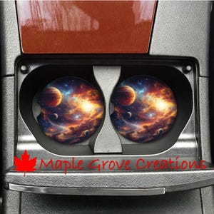 Peut inclure: Deux dessous de verre pour voiture avec un design de galaxie vibrant, représentant des planètes, des étoiles et des nébuleuses dans des tons orange, bleu et violet. Les dessous de verre sont ronds et s'insèrent dans les porte-gobelets d'une voiture. Le texte "Maple Grove Creations" est visible.
