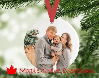 Peut inclure: Une boule de Noël ronde, blanche et en céramique avec une photo d'une famille de trois personnes. La photo montre un homme, une femme et un jeune enfant. L'homme porte un pull gris et la femme porte une chemise grise. L'enfant porte une chemise blanche. La boule de Noël est munie d'un ruban rouge.