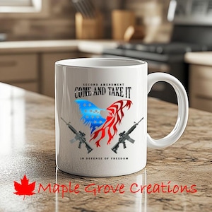 Pode incluir: Caneca de café de cerâmica branca com um design de bandeira americana vermelha, branca e azul. A caneca tem o texto "Second Amendment Come and Take It" e "In Defense of Freedom".