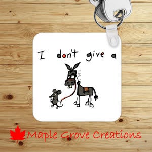 May include: White square keychain with the text "I don't give a" and a cartoon donkey being led by a mouse. The keychain has a silver key ring and a red maple leaf logo with the words "Maple Grove Creations".