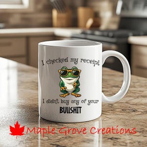 Może przedstawiać: Biały ceramiczny kubek z żabą w okularach przeciwsłonecznych i napisem "I checked my receipt, I didn't buy any of your BULLSHIT." Kubek ma również napis "Maple Grove Creations" i czerwony liść klonu.