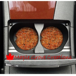 May include: Two round brick-patterned car coasters in a black cup holder. The coasters are made of a durable material and are designed to protect your car's cup holder from spills and scratches. The coasters feature a realistic brick pattern with a distressed look. The text "Maple Grove Creations" is visible below the cup holder.