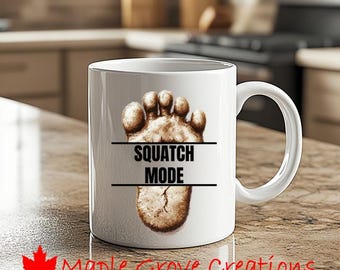 May include: White ceramic mug featuring a brown Bigfoot footprint design with the text "SQUATCH MODE" in black. The mug has a curved handle and is suitable for hot or cold beverages. The Maple Grove Creations logo is at the bottom.