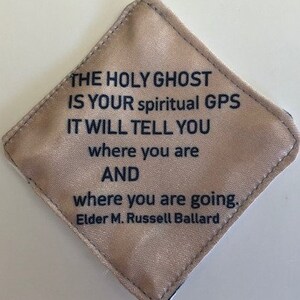 May include: A diamond-shaped, beige fabric item with black text. The text reads "THE HOLY GHOST IS YOUR spiritual GPS IT WILL TELL YOU where you are AND where you are going. Elder M. Russell Ballard."