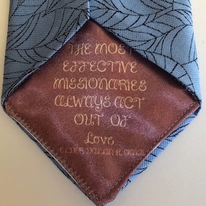 May include: A blue tie with a leaf pattern and a pink lining. The lining has the text "The most effective missionaries always act out of love."
