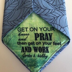 May include: A blue tie with a white leaf pattern. The tie is flipped to show the inside lining, which is green with a quote that reads "Get on your knees and pray, then get on your feet and work. Gordon b. hinkley."