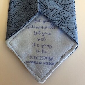 May include: A blue tie with a leaf pattern. The tie has a white lining with the text "Eat your vitamin pills Get your rest It's going to be EXCITING! RUSSELL M. NELSON" printed on it.