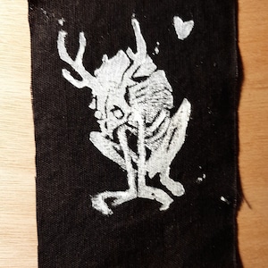 Op de afbeelding: Een zwarte stoffen patch met een witte omtrek van een skelettig hert met grote geweien. Het hert zit met zijn hoofd gebogen. Een klein wit hartje bevindt zich in de rechterbovenhoek van de patch.