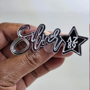 May include: Silver acrylic pin with the word "Silver" in a cursive font and a black star with the letters "AZ 89" inside.