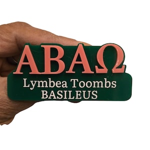 May include: A green and peach-colored name tag with the Greek letters ABAΩ. The name tag also includes the name "Lymbea Toombs" and the word "BASILEUS" in white text.