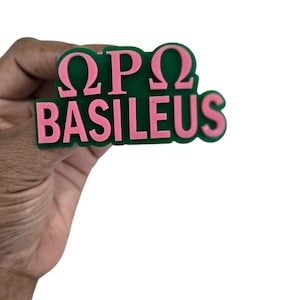 May include: A green and pink plastic pin with the letters "ΩΡΩ" and the word "BASILEUS" in pink.