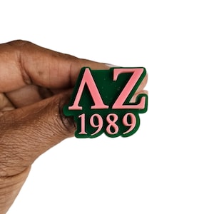 May include: A green and pink plastic pin with the letters "AZ" in pink and the year "1989" in pink below.