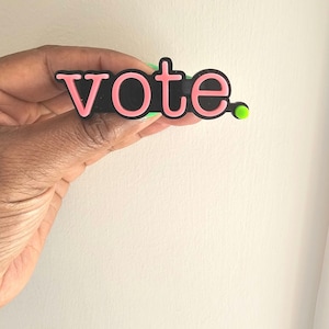 May include: A black and pink plastic pin with the word "vote" in pink letters. The pin has a green dot on the right side.