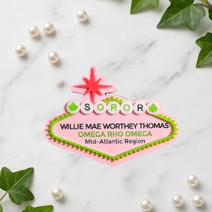 May include: A pink and green custom name badge shaped like a retro sign. It features a red star at the top, white circular accents with green ivy leaves, and the text: SOROR, WILLIE MAE WORTHEY THOMAS, OMEGA RHO OMEGA, and Mid-Atlantic Region.