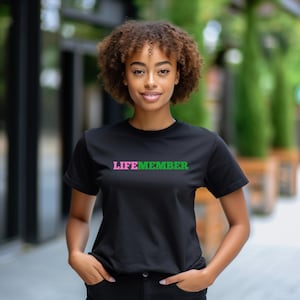May include: Black t-shirt with the word "LIFEMEMBER" printed across the chest in pink and green. The shirt is a crew neck style and has short sleeves. The model is wearing black pants.