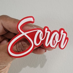 May include: Red and white acrylic word "soror" with a white outline. The word is in a cursive font.