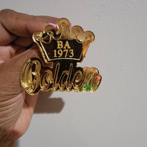 May include: Gold mirror acrylic ornament with a crown and the text "BA 1973 Golden".