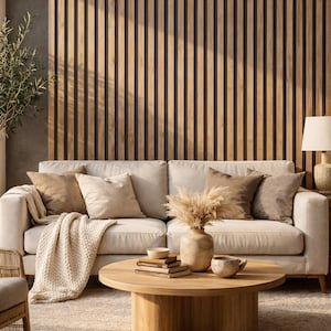 May include: A living room scene featuring a beige sofa with several throw pillows and a knitted blanket. A round wooden coffee table holds books, a vase with dried flowers, and ceramic bowls. The background includes a wooden slatted wall.