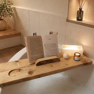 May include: A natural wood bathtub tray resting across a white tub filled with bubble bath. The tray features a central wooden book stand holding an open book, a circular cutout, a small white sphere, a rectangular light, and a small amber glass candle.