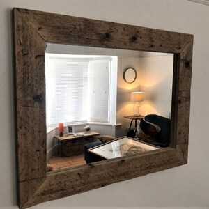Op de afbeelding: Een grote rechthoekige spiegel met een rustieke houten lijst. De lijst is gemaakt van gerecycled hout met een natuurlijke bruine afwerking. De spiegel reflecteert een woonkamer met een raam, een lamp en een bank.