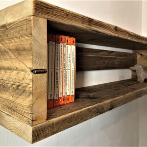 May include: Rustic wooden shelf with a unique angled design. The shelf has a natural wood finish and features a small wooden dachshund figurine on the lower shelf. A row of orange paperback books are displayed on the upper shelf.