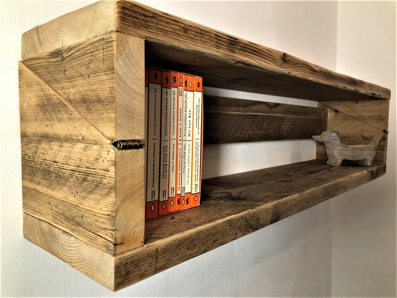 May include: Rustic wooden shelf with a unique angled design. The shelf has a natural wood finish and features a small wooden dachshund figurine on the lower shelf. A row of orange paperback books are displayed on the upper shelf.