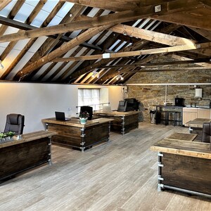 May include: A modern office space with rustic wooden desks and exposed beams. The desks are made of reclaimed wood and have industrial metal legs. The room has a large window and a brick wall.