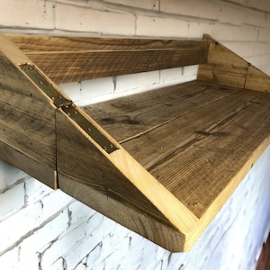 May include: A rustic wooden shelf with a triangular design and brass hinges. The shelf is mounted to a white brick wall.