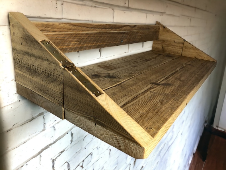 May include: A rustic wooden shelf with a triangular design and brass hinges. The shelf is mounted to a white brick wall.