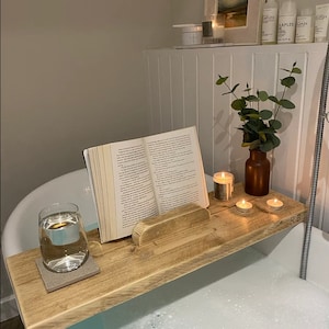 Puede incluir: Una bandeja de baño de madera con un soporte para libros, dos velas y un vaso de agua. La bandeja está colocada en el borde de una bañera llena de burbujas. La bandeja está hecha de madera reciclada y tiene un aspecto rústico.