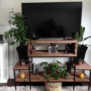 May include: A black metal shelf unit with three tiers, featuring a wooden top shelf and two lower shelves. The unit is decorated with plants, decorative objects, and a flat screen TV.