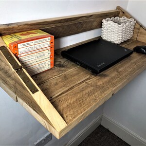 May include: A rustic wooden wall-mounted desk with a laptop, a white woven basket, and a computer mouse. The desk is made of reclaimed wood and has a natural finish. The desk is mounted to the wall with hinges.