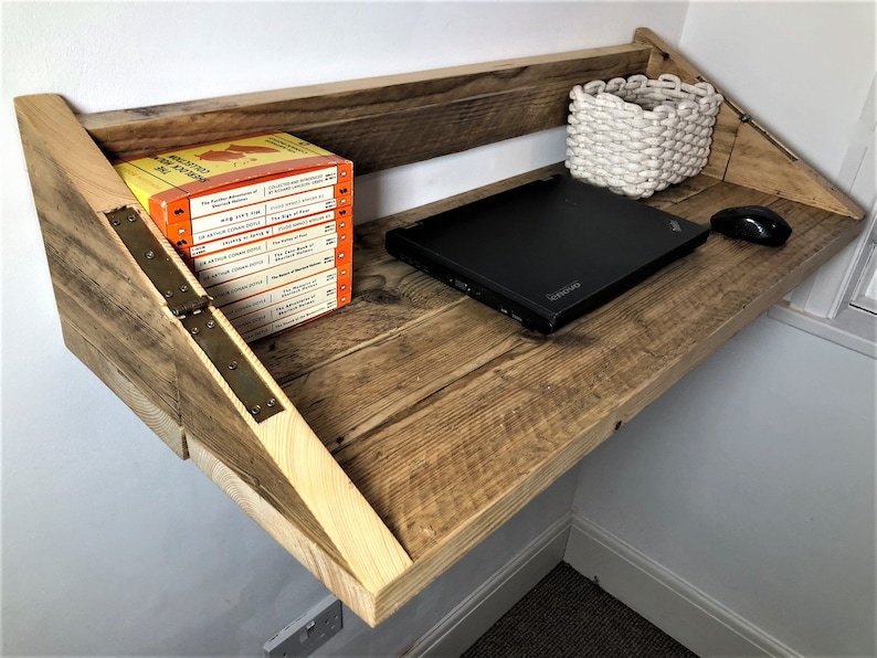 May include: A rustic wooden wall-mounted desk with a laptop, a white woven basket, and a computer mouse. The desk is made of reclaimed wood and has a natural finish. The desk is mounted to the wall with hinges.