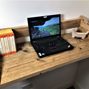 May include: A rustic wooden wall-mounted desk with a laptop, a stack of books, and a wooden dog figurine. The desk is made of reclaimed wood and has a natural finish. The laptop is open and has a screen displaying the time and date. The books are stacked neatly on a shelf. The dog figurine is sitting on the desk.