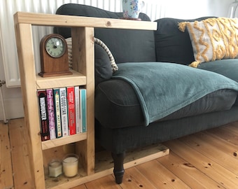 May include: A wooden side table with a shelf for books and a clock. The table is next to a gray couch with a throw blanket and a pillow.