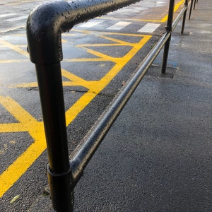 May include: Black metal railing with a black pipe and a yellow painted crosswalk in the background. The railing is wet from rain and has water droplets on it.