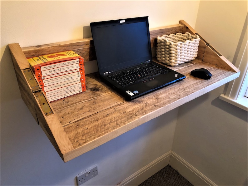 May include: A wooden wall-mounted desk with a laptop, a white woven basket, and a computer mouse. The desk is made of reclaimed wood and has a natural finish. There are several books stacked on the left side of the desk.