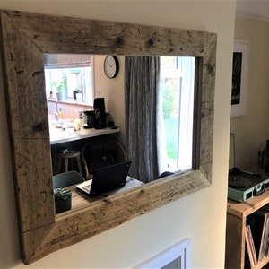 Op de afbeelding: Een grote rechthoekige spiegel met een rustieke houten lijst. De lijst is gemaakt van gerecycled hout met knopen en onvolkomenheden. De spiegel reflecteert een kamer met een raam, een tafel en een laptop.