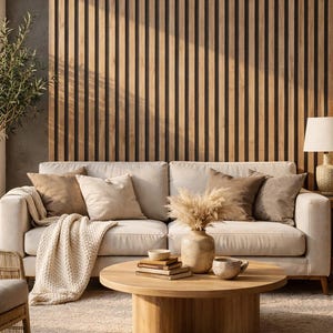 May include: A cozy living room scene featuring a cream-colored sofa with several throw pillows and a knitted blanket. A round wooden coffee table holds books, a vase with dried flowers, and ceramic bowls. The background has a wooden slat wall.