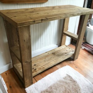 May include: A rustic wooden console table with two shelves. The table is made of reclaimed wood and has a natural finish. The table is 76 cm tall and 91 cm wide.