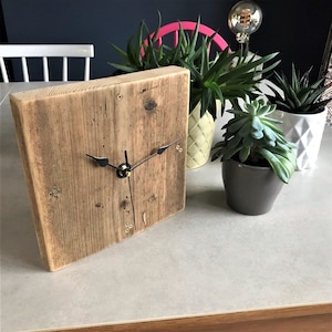 Puede incluir: Un reloj de madera rústico con manecillas negras y detalles dorados. El reloj está sobre una superficie gris claro con plantas en macetas en el fondo.