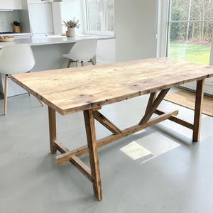 Pode incluir: Uma mesa de jantar retangular feita de madeira castanho-clara com um padrão de grão visível. A mesa apresenta uma superfície superior espessa e plana, suportada por quatro pernas robustas ligadas por uma viga central e reforços diagonais.