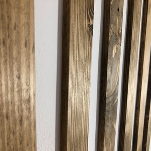 May include: Close-up of vertical wooden slats with a natural, weathered finish. The wood grain is visible, and the slats are arranged against a white background. The varying tones of brown and gray create a rustic aesthetic.