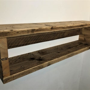 May include: Rustic wooden shelf with a natural finish. The shelf has a rectangular shape and is made of reclaimed wood. It has a single open shelf and is mounted to a wall.