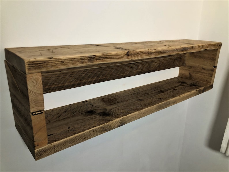 May include: Rustic wooden shelf with a natural finish. The shelf has a rectangular shape and is made of reclaimed wood. It has a single open shelf and is mounted to a wall.